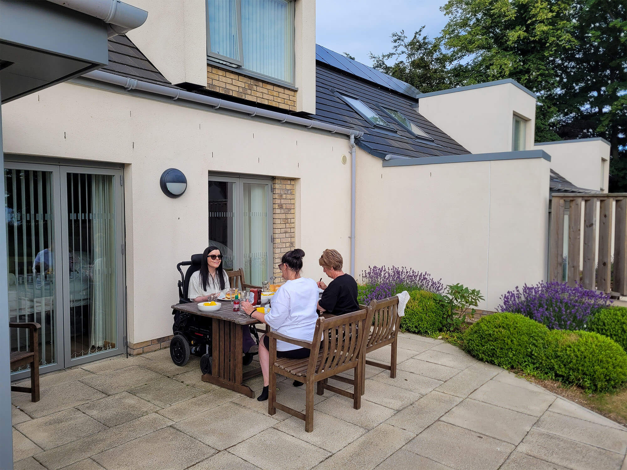 Emma is with her sister and mum sitting on the patio at the table eating BBQ food.
