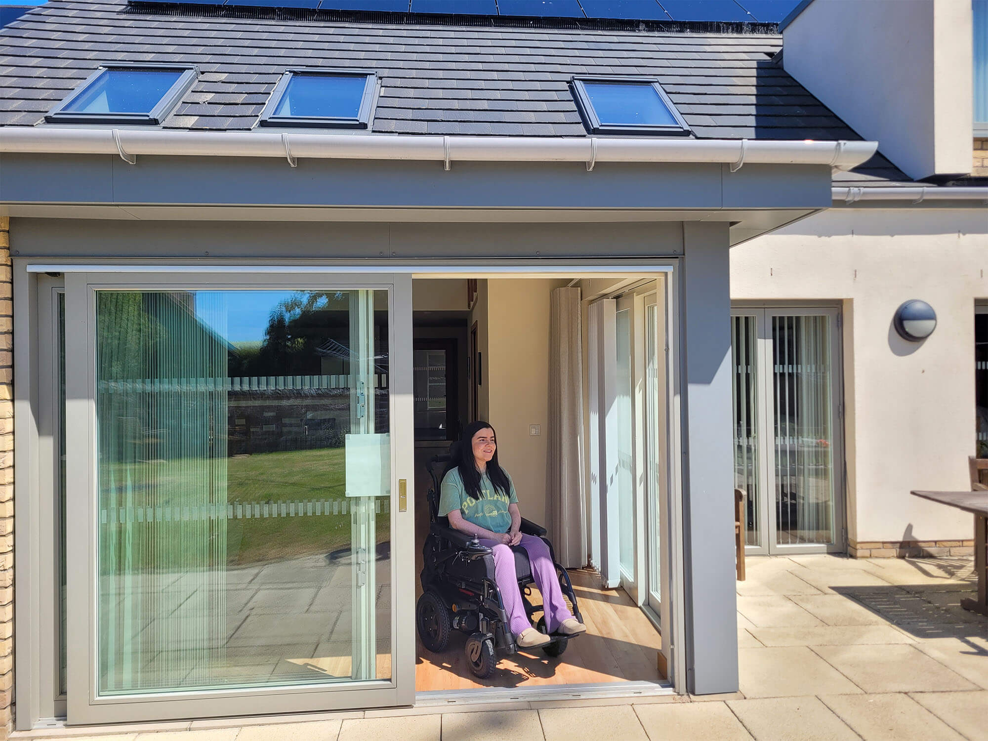 Emma in her wheelchair sat inside the patio doors of the Largo lodge at Homelands Trust-Fife. Emma is wearing a green t-shirt and purple trousers.