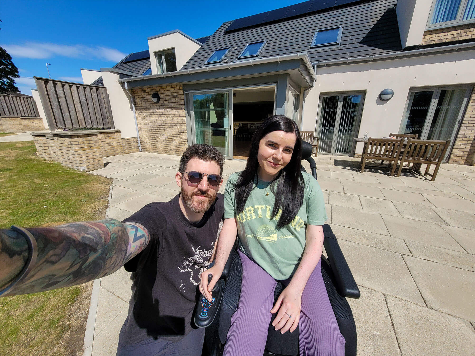 Emma in her wheelchair and her partner, sat outside in front of the Largo lodge at Homelands Trust-Fife, a wheelchair accessible lodge in Scotland. Emma is wearing a green t-shirt and purple trousers and her partner is wearing a black t-shirt and sunglasses.