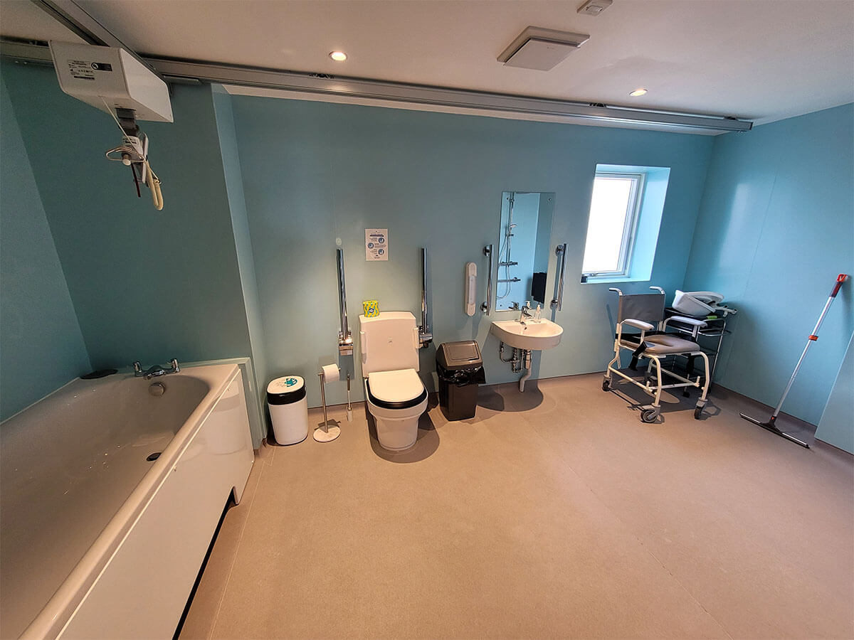 A large wet room in the Largo accessible lodge at Homelands Trust. It has a clos-o-mat toilet, shower with wall mounted seat, shower commode chair, accessible bath and ceiling track hoist.
