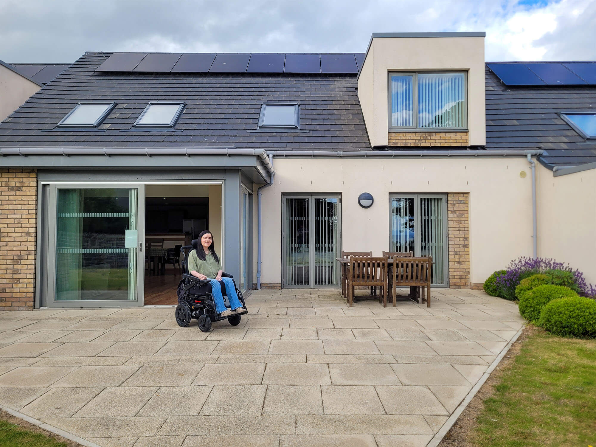 Emma in her wheelchair sat outside in front of the Largo lodge at Homelands Trust-Fife, a wheelchair accessible lodge in Scotland. Emma is wearing a green shirt and jeans.