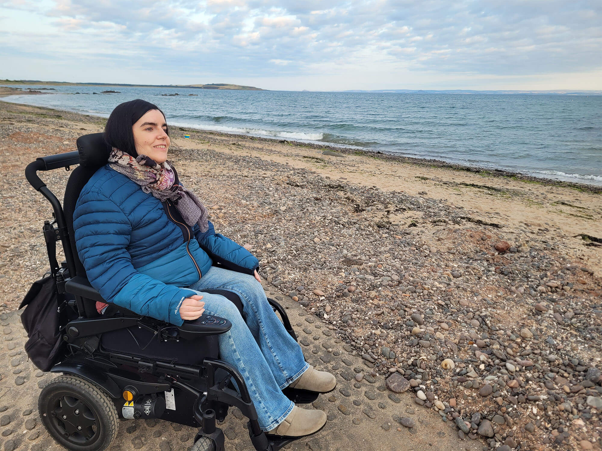 Emma, a wheelchair user is on Lower Largo Beach. Emma is smiling and looking at the sea. She is wearing a puffer jacket, jeans and a scarf.