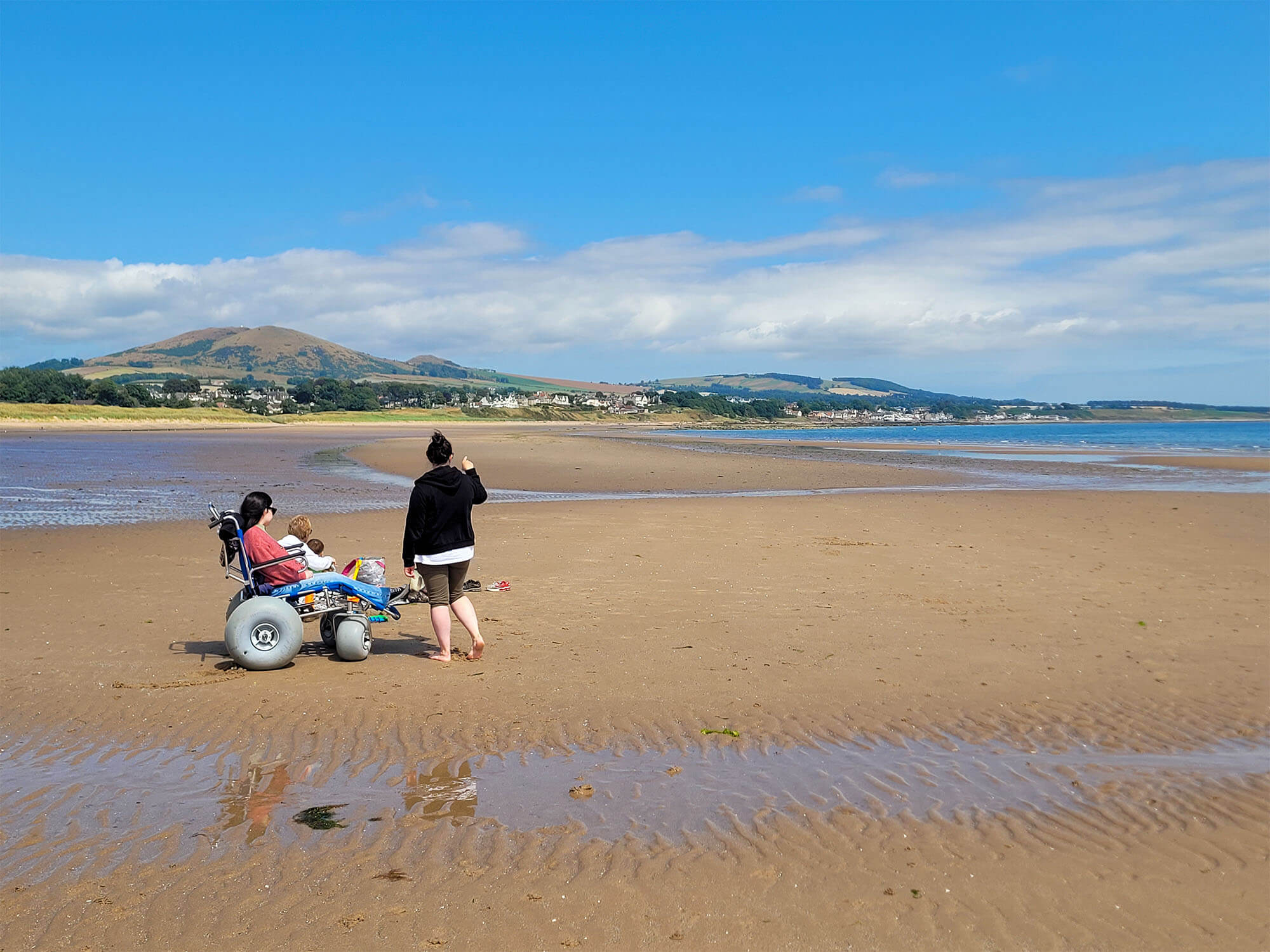 Emma sat in a manual sandcruiser with balloon wheels from Silverburn Park Beach Wheelchairs. She is sat with her family on the beach. Hills can be seen in the distance.
