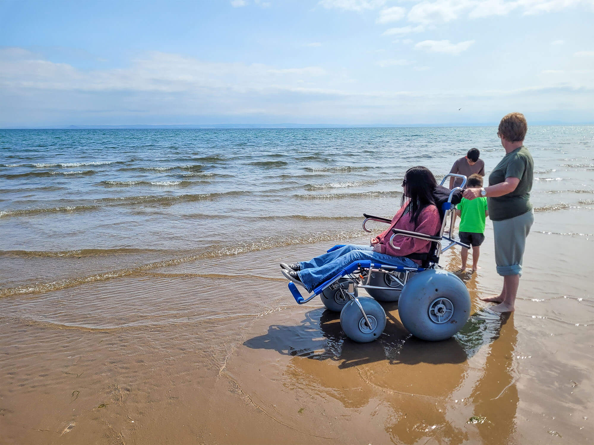 Emma is sat in a beach wheelchair on Silverburn Park beach. She is being pushed by her mum and her two young nephews are walking barefoot alongside them.