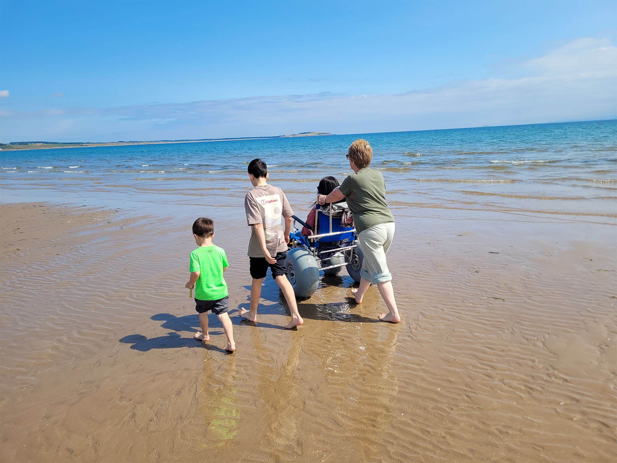 Emma is sat in a beach wheelchair on Silverburn Park beach. She is being pushed by her mum and her two young nephews are walking barefoot alongside them.