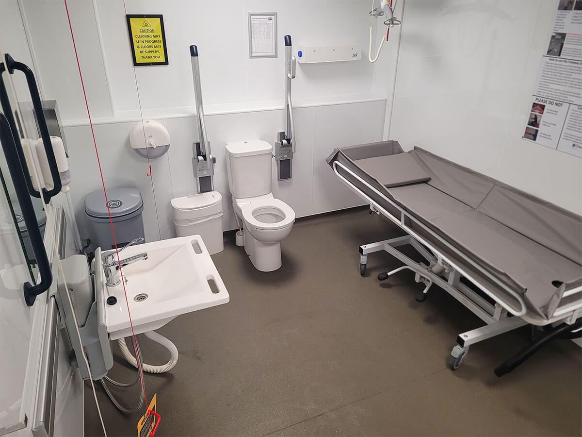 Interior of a Changing Places toilet showing the adult-sized changing bed, peninsular toilet and sink, ceiling-track hoist, grab rails and a shower.