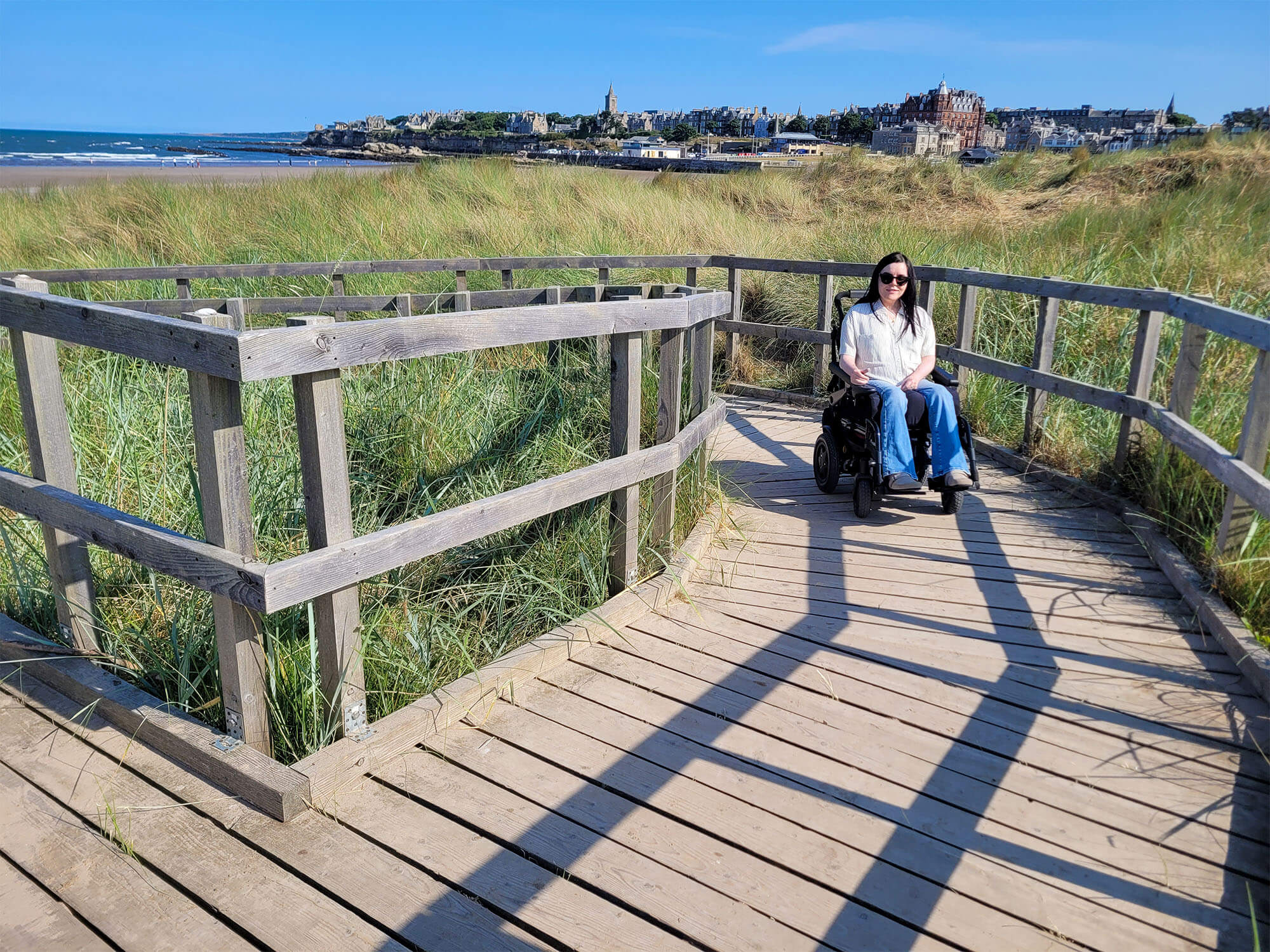 Emma, a wheelchair user is on the wooden boardwalk at St Andrews West Sands Beach. Emma is smiling at the camera. It is a sunny day.