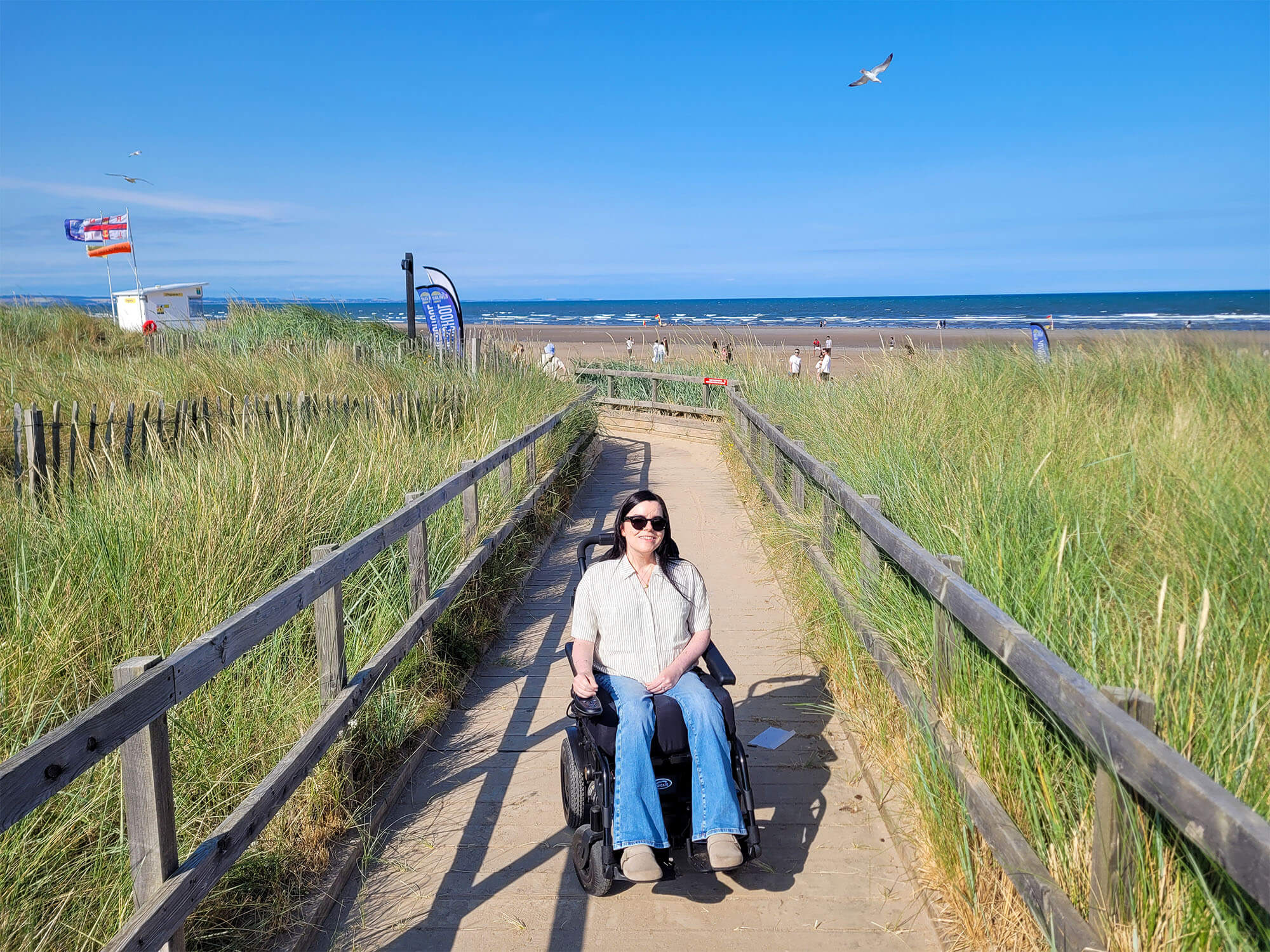 Emma, a wheelchair user is on the wooden boardwalk at St Andrews West Sands Beach. Emma is smiling at the camera. It is a sunny day. People are on the beach behind Emma and a seagull is in the sky.
