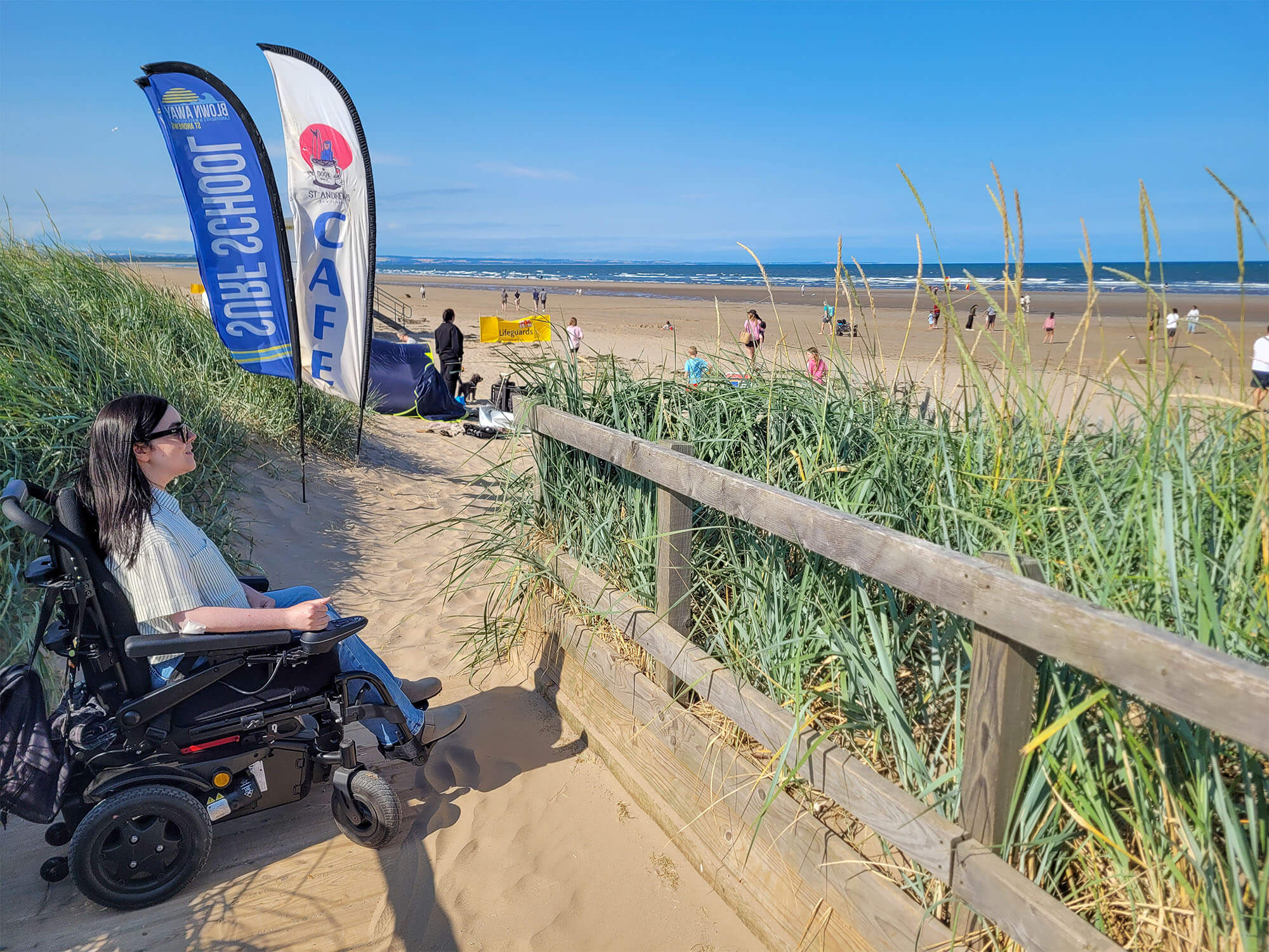 Emma, a wheelchair user is on the wooden boardwalk at St Andrews West Sands Beach. Emma is smiling as she looks out to the sea. It is a sunny day. People are on the beach.