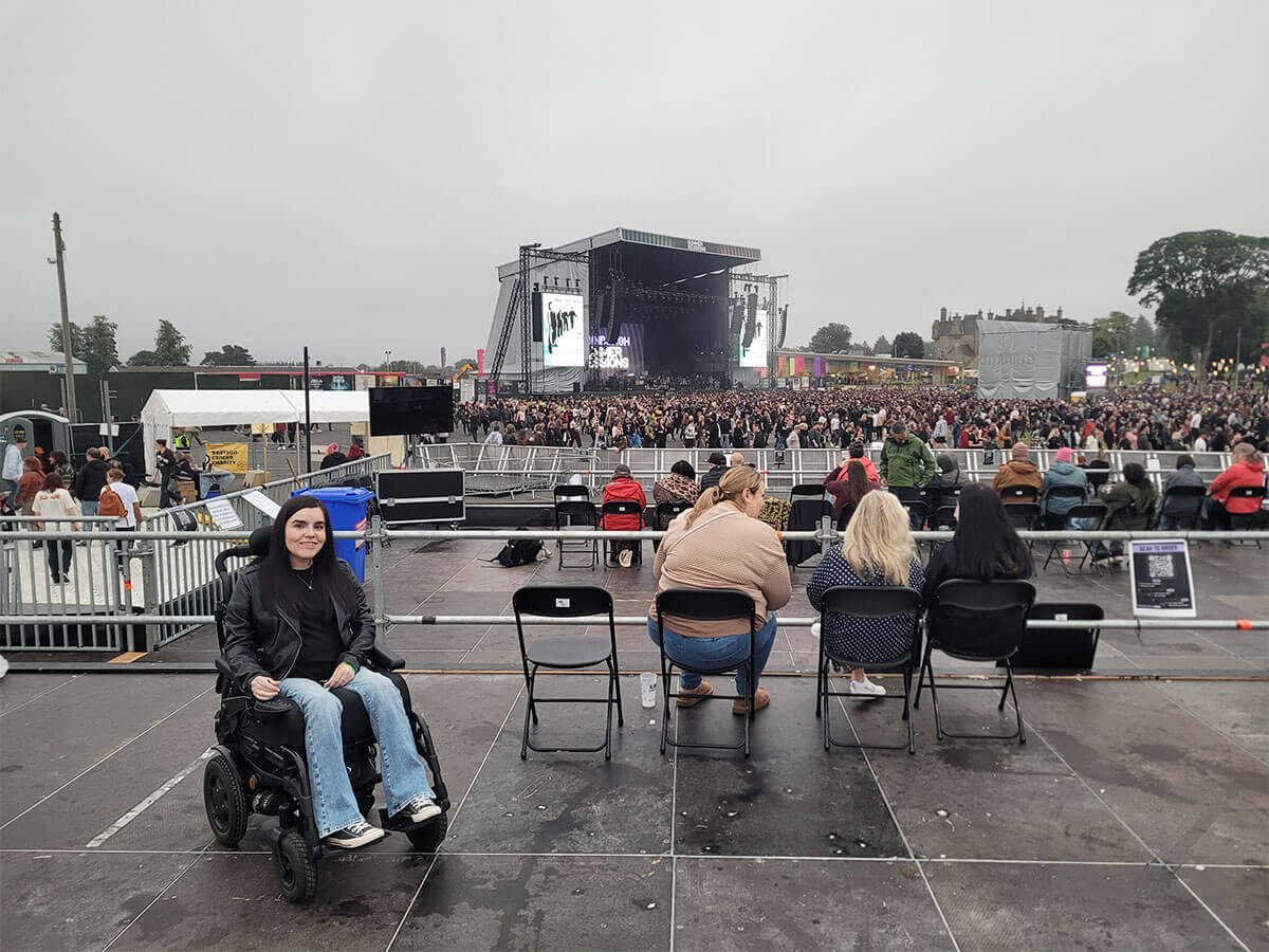 Emma sat on the Edinburgh Summer Sessions accessible viewing platform. She is smiling at the camera. The crowd and stage are behind Emma.