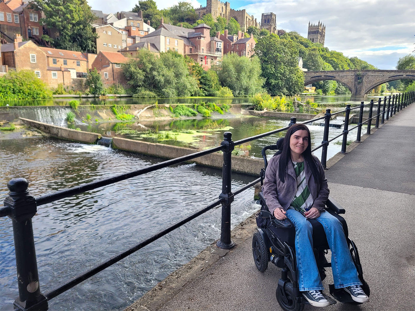 Emma, sitting in her wheelchair alongside the river wear with Durham Castle and Cathedral in the background.