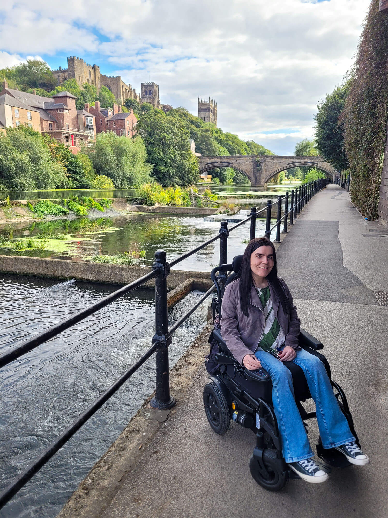 Emma, sitting in her wheelchair alongside the river wear with Durham Castle and Cathedral in the background.