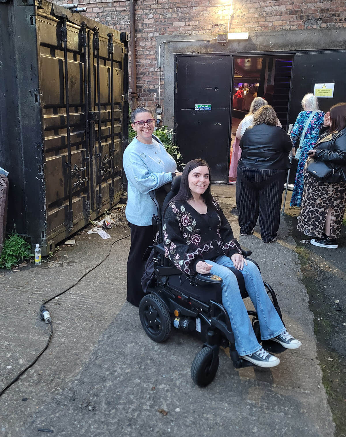Emma, a female wheelchair user, and her sister are outside of The Garage at the wheelchair accessible entrance. They are both smiling at the camera.
