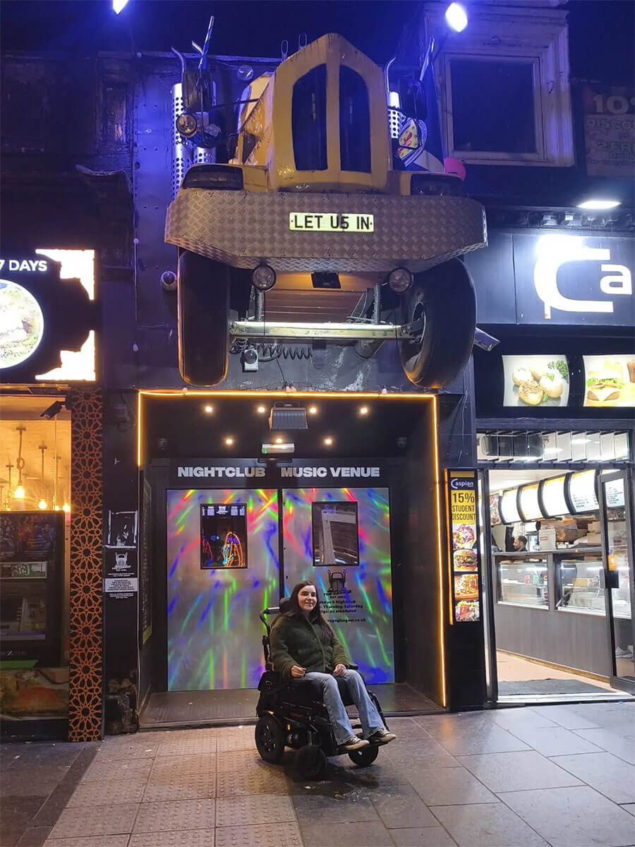 Emma sat in her wheelchair outside of The Garage nightclub. She is sat in front of the main entrance below the iconic yellow truck coming out of the building.