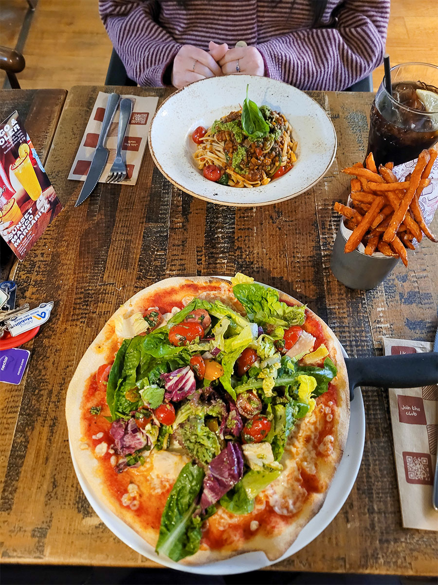 Two plates on a table. One with pizza and one with pasta plus a portion of sweet potato fries and a glass of diet coke.